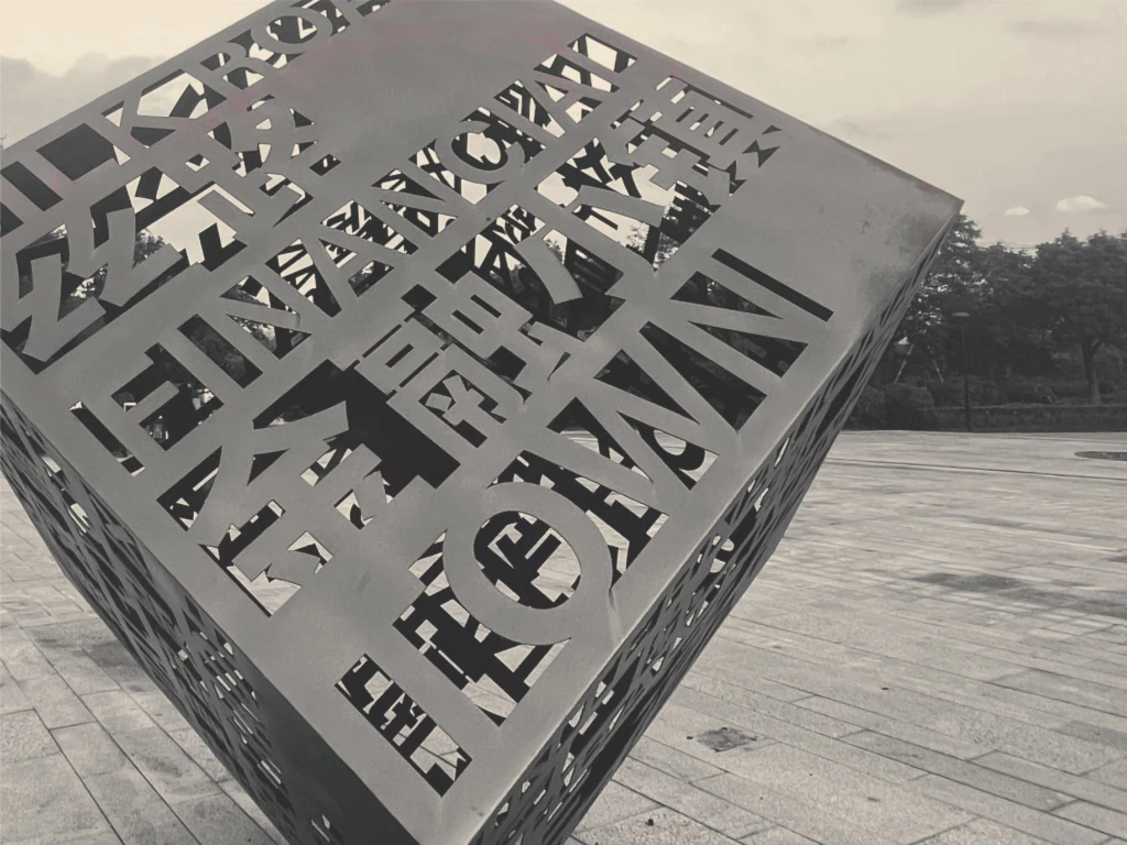 Yiwu- Imagen monocromática de una gran escultura cúbica con intrincadas letras recortadas, expuesta al aire libre en un área pavimentada.