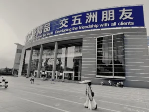 Yiwu- Un gran edificio con un cartel que dice "Desarrollando la amistad con todos los clientes". Varias personas caminan frente al edificio y una de ellas sostiene un paraguas.